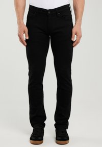 Mann trägt schmal geschnittene schwarze Jeans und schwarze Sneaker mit Gummisohlen, steht vor einem schlichten, hellen Hintergrund.