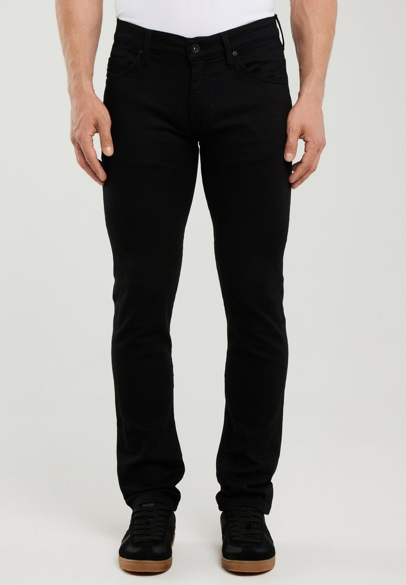 Mann trägt schmal geschnittene schwarze Jeans und schwarze Sneaker mit Gummisohlen, steht vor einem schlichten, hellen Hintergrund.