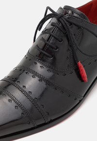 Scarpa in pelle nera con motivo a diamante in rilievo, punta rotonda e lacci neri; presenta dettagli rossi vicino ai passanti.