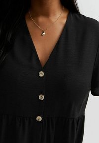 Woman wearing a black V-neck dress with three beige buttons and a gold chain necklace with a small pendant.