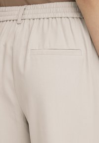 Pantalon beige en tissu doux et texturé. Comprend une ceinture élastique et une poche arrière unique avec un détail de couture subtil.