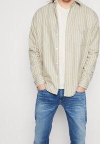 Camisa a rayas beige claro con botón en el frente y un bolsillo en el pecho, combinada con jeans azules. Tejido de algodón suave, corte relajado.