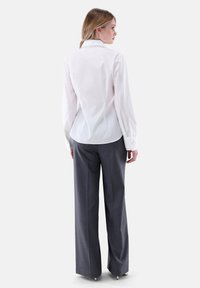 Camicia bianca a maniche lunghe con orlo curvo e dettagli decorativi nella collare, abbinata a pantaloni grigi a gamba larga, che mostrano una texture liscia.