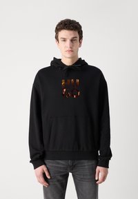 Sudadera negra con bolsillo canguro, capucha con cordón y un brillante logo "HUGO" en naranja y dorado impreso en la parte delantera.