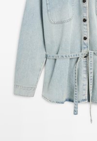 Veste en jean bleu clair à manches longues, boutons noirs sur le devant, grande poche poitrine et ceinture assortie nouée à la taille.