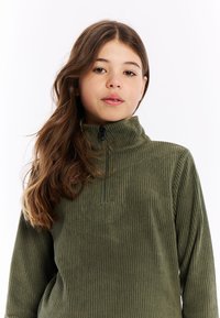 Meisje met lang bruin haar, gekleed in een olijfgroene ribbeltrui met een halve ritssluiting, recht vooruit kijkend tegen een witte achtergrond.