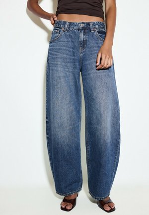 Person trägt hoch taillierte, weit geschnittene blaue Jeans mit verwaschenem Look, schwarzen kurzen Top und dunkle offene Sandalen mit Absatz vor weißem Hintergrund.