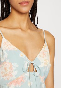 Vestido floral azul claro com alças finas ajustáveis, apresentando um recorte em forma de chave e um detalhe de laço na gola. Tecido suave e texturizado.