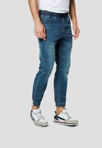 Spodnie dresowe z denimu w ciemnym niebieskim kolorze z elastycznym paskiem i ściągaczami, z kontrastowym przeszyciem paneli i wieloma kieszeniami.