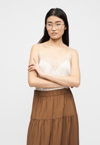 Νεαρή γυναίκα με διαφανή γυαλιά, λευκό τοπ με δαντέλα και καφέ φούστα με στρώσεις, στέκεται με σταυρωμένα τα χέρια μπροστά σε λευκό φόντο.