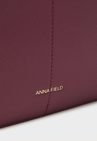 Bordeaux teksturert veske med glatt finish, med gull "ANNA FIELD" merke og rene sømdetaljer.
