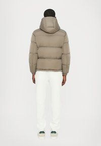 Puffer jacket in muted taupe with a hood, featuring quilted panels. Paired with white sweatpants and grey-accented sneakers.