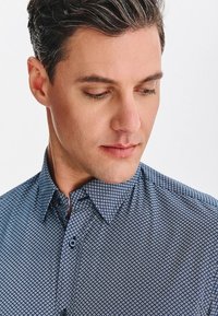 Niebieska koszula w wzory o geometrycznym wzorze, zapinana na guziki z standardowym kołnierzykiem. Materiał wydaje się lekki o gładkiej teksturze.