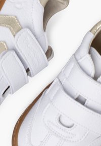 Bílé syntetické boty se zlatými akcenty, vybavené dvěma suchými zipy a hnědou gumovou podrážkou, zdůrazňující elegantní design a strukturovaný povrch.