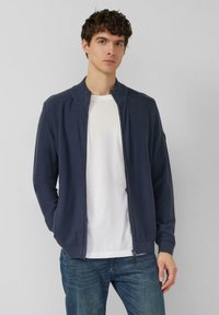 Marineblauwe vest met ritssluiting van gladgebreide stof, voorzien van een geribde kraag en manchetten, gecombineerd met een wit T-shirt en blauwe jeans.