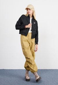 Schwarze, verkürzte Bomberjacke mit gerippten Bündchen, weißem Logo, beigem Hut, olivgrünen Cargo-Hosen mit Seitentaschen und sandfarbenen Schnürstiefeln mit dicken Sohlen.
