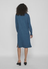 Kvinna med lockigt hår, klädd i en knälång blå stickad klänning och svarta klackskor, stående med ryggen mot en enkel bakgrund.