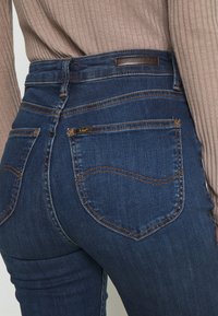 Tmavě modré džíny s vysokým pasem a slim fit, s kontrastním prošíváním, dvěma zadními kapsami a malou logovou nášivkou na pasu.