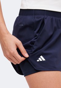 Main ajustant un short de sport Adidas ClimaCool bleu marine sur une couche intérieure noire, portant un haut blanc.