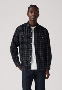Black and gray plaid shirt with two chest pockets, white buttons, and a relaxed fit, layered over a white T-shirt.