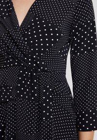 Robe portefeuille noire avec des pois blancs variés et des manches trois-quarts, nouée à la taille avec une ceinture en tissu.