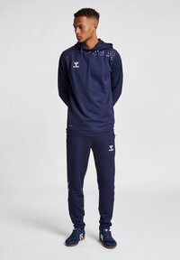 Marineblaues Sporttracksuit mit Kapuze, das ein gepunktetes Muster auf den Schultern und kontrastierende Streifen an den Hosenbeinen aufweist. Enthält Logodetails.