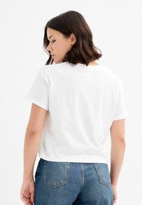 Camiseta blanca de algodón con mangas cortas, cuello redondo y corte relajado, combinada con jeans de denim azul que cuentan con bolsillos traseros.