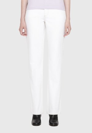 Person wearing straight-cut white pants, light top, and black ankle boots, standing against a plain light gray background.