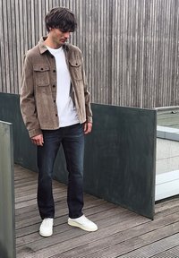 Mann trägt braune Cordjacke, weißes T-Shirt, dunkle Jeans und weiße Sneakers, steht auf einem Holzdeck neben grauen Paneelen und einem vertikalen Holzzaun.