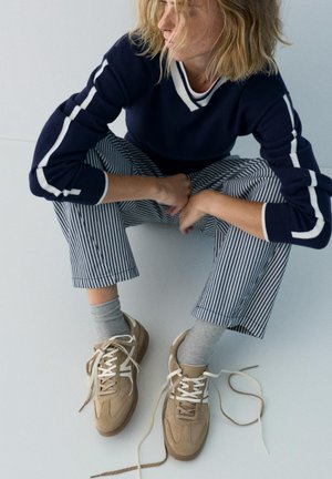 Osoba o blond włosach siedząca po turecku, ubrana w granatowy sweter w białe paski, spodnie w paski, szare skarpetki i beżowe trampki ze związanymi na luźno sznurówkami.