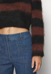 Personne portant un pull court à rayures noires et brunes en matière duveteuse et un jean bleu taille haute avec poches plaquées à l'avant, main détendue le long du corps.