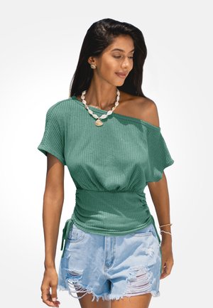 Vrouw draagt groene off-shoulder geribde top, versleten lichtblauwe denim shorts, schelpenketting en schelpvormige oorbellen tegen witte achtergrond.