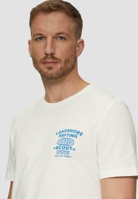 Witte katoenen t-shirt met een blauwe grafische print met de tekst "LAKESHORE RAFTING" en "SCOUT HARDWARE & SUPPLIES" op de borst. Korte mouwen.