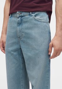Homme portant un jean denim bleu clair et une chemise bordeaux foncé, visible de la taille jusqu'au milieu de la cuisse, les mains détendues le long du corps.