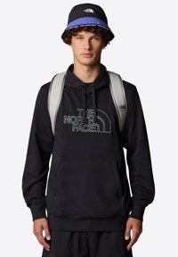 Felpa nera con tasca frontale e logo a contrasto, spallacci dello zaino grigi, cappello a secchiello nero con bordo blu, indossato sopra pantaloni neri.