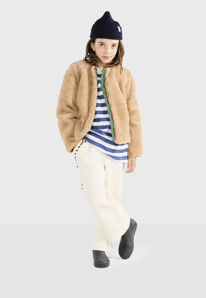 Tan faux fur jakke, marineblå strikket hue, blå og hvid stribet skjorte, cremefarvede vidde bukser med sorte accenter, sort fodtøj.