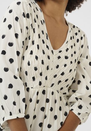Blouse blanche à pois noirs, décolleté en v, détails smockés et manches bouffantes avec des poignets élastiques, créant une apparence texturée.