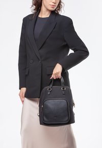 Borsa nera in materiale sintetico con una tasca frontale testurizzata, manici superiori doppi e minuteria in metallo dorato; abbinata a un blazer nero e una gonna color crema.