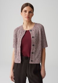 Kurzärmlige, kastanienbraune und weiße Strickjacke mit Rundhalsausschnitt, fünf schwarzen Knöpfen und einem strukturierten Muster. Kombiniert mit einem kastanienbraunen Oberteil.