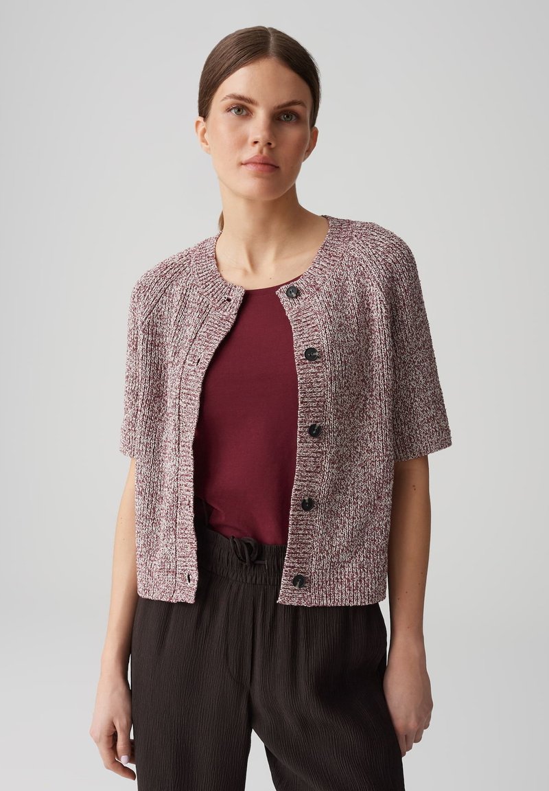 Kurzärmlige, kastanienbraune und weiße Strickjacke mit Rundhalsausschnitt, fünf schwarzen Knöpfen und einem strukturierten Muster. Kombiniert mit einem kastanienbraunen Oberteil.