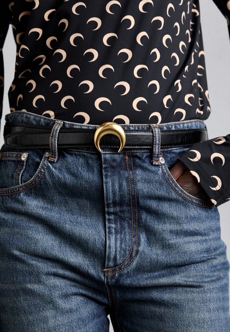 Black leather belt with a gold crescent-shaped buckle worn over blue denim jeans and patterned black top featuring beige crescent shapes.