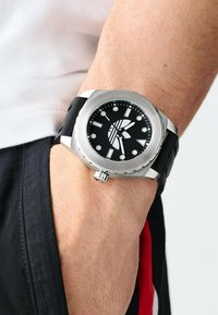 Montre avec bracelet en caoutchouc noir, présentant un boîtier rond en acier argenté, un cadran noir avec des index et aiguilles blancs, une couronne surdimensionnée et un design épuré.