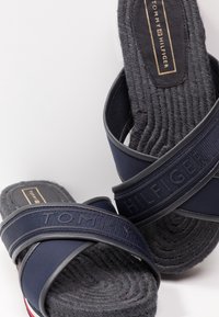 Sandales mules bleu marine avec des brides en tissu texturé, affichant le nom de la marque en relief ; semelle tissée noire avec un motif circulaire.