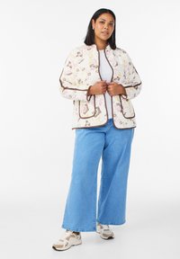 Gewatteerd jack met bloemen- en paisleypatronen in crèmekleur, afgewerkt met bruine bies. Gecombineerd met lichtblauwe jeans met wijde pijpen en beige sneakers.