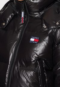Veste matelassée noire brillante avec fermeture éclair devant, poches à boutons-pression et écusson avec logo Tommy Jeans sur la poitrine.