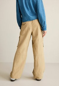 Beige vidde cargo bukser med sidelommer og afslappet pasform, kombineret med en blå denim skjorte med lange ærmer.