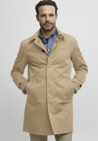 Manteau beige en trench avec poches à boutonnière, texture douce et design classique. Il présente un petit logo sur la poitrine et un col.