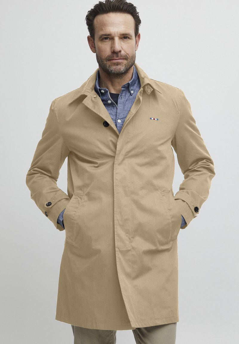 Manteau beige en trench avec poches à boutonnière, texture douce et design classique. Il présente un petit logo sur la poitrine et un col.
