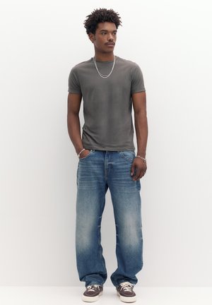 Homme aux cheveux courts et bouclés portant un t-shirt gris, un jean bleu ample, des baskets, ainsi qu'une chaîne et des bracelets en argent, debout les mains dans les poches.