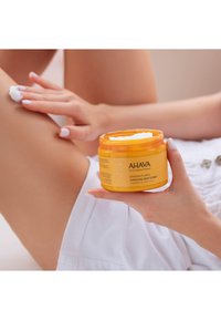 Gul burk med AHAVA Dead Sea Plants Caressing Body Sorbet, hålls i en hand, med vit kräm synlig på toppen och applicerad på ett ben.